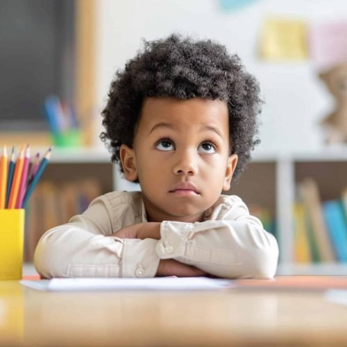 A young boy sits at a desk with a pencil and a piece of paper in front of him. He looks sad and is staring at the paper